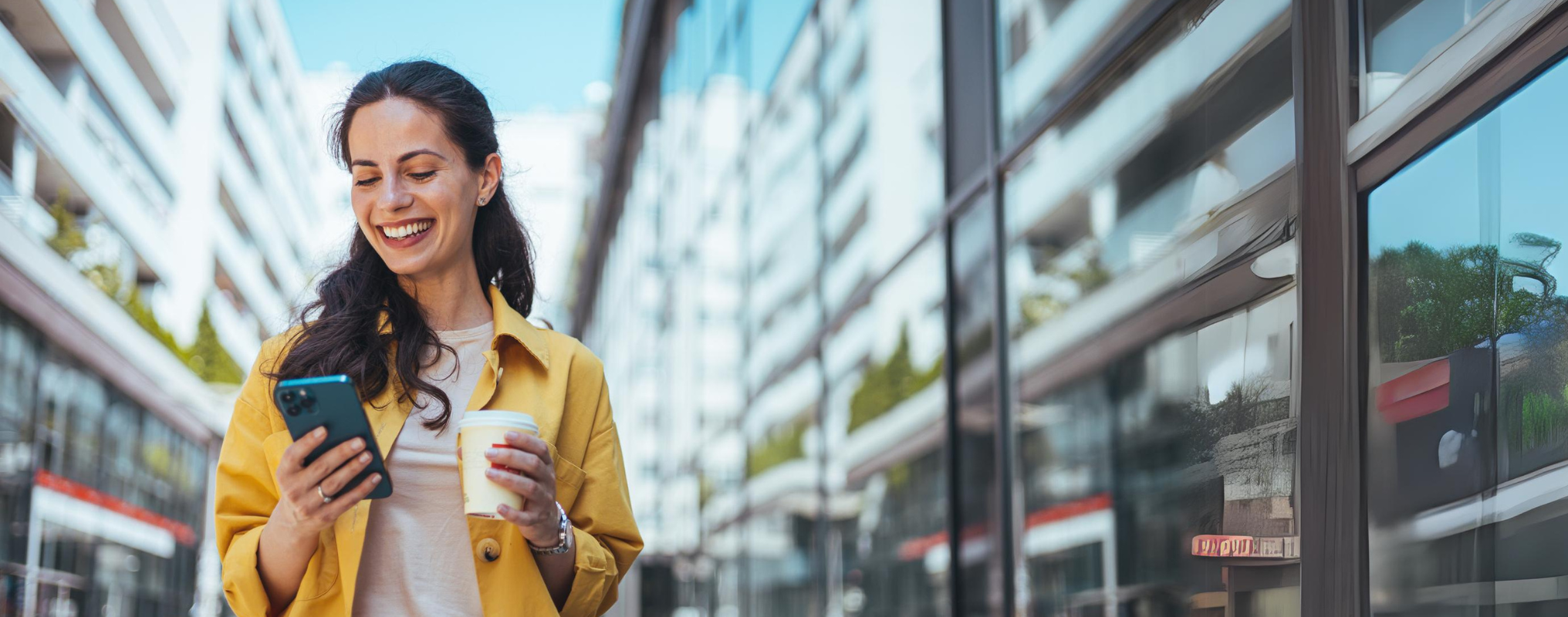 Eine Frau läuft lächelnd durch die Straße, hält in einer Hand einen Kaffee, in der anderen ihr Handy